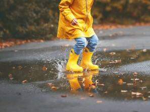 A child in a yellow raincoat and boots is splashing in a puddle
